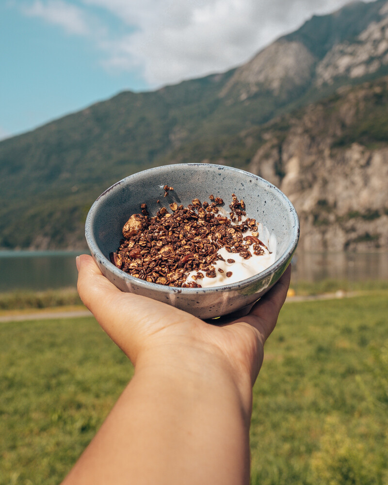 Crunchy chocolate muesli with yogurt in bowl in front of mountain and lake.